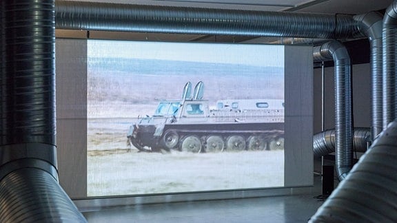 Große, metallische Rohre winden sich in einem Ausstellungraum um eine Videoleinwand.