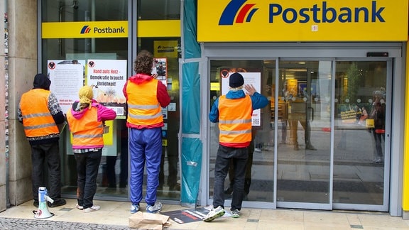 Vier Personen kleben Plakate auf eine Postbankfiliale.