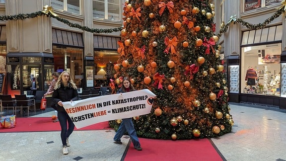 Zwei Frauen halten ein Plakat vor einem mit Farbe besprühten Weihnachtsbaum.