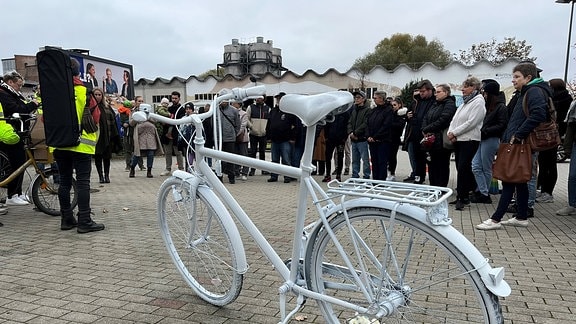 Menschen stehen auf einem Supermarktparkplatz. Im Vordergrund ein weißes Fahrrad