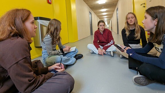 Fünf Schülerinnen sitzen in einem Schulflur und diskutieren miteinander. 