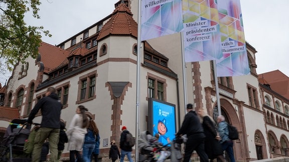 Menschen überqueren vor der Leipziger Kongresshalle und wehenden Fahnen die Straße.