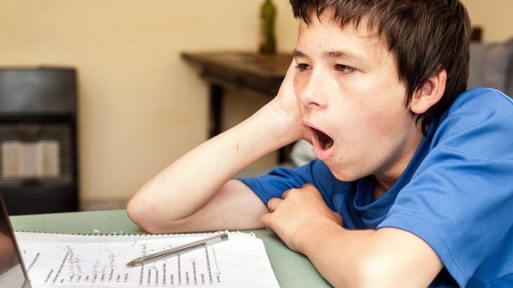 Müder Schüler sitzt gähnend über den Hausaufgaben