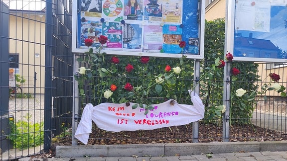 Rosen und Banner hängen an einem Zaun bei einer Gedenkveranstaltung für einen Ermordeten. 