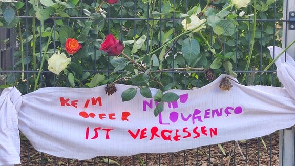 Rosen und Banner hängen an einem Zaun bei einer Gedenkveranstaltung für einen Ermordeten. 