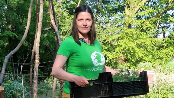Eine Frau mit grünem T-Shirt steht mit Korb in einem Garten