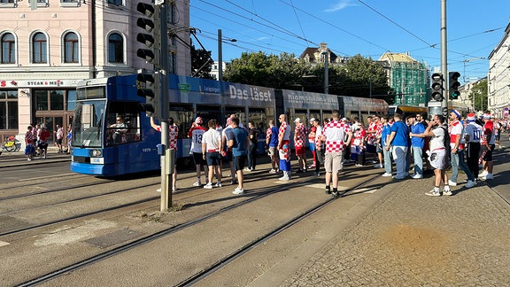An einer Straßenbahnhaltestelle queren Zahlreiche Menschen mit Trikots der kroatischen Nationalmannschaft die Schienen. Im Hintergrund fährt eine Straßenbahn.