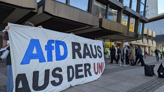 Vor der Universität stehen junge Menschen und halten ein Tranpsarant hoch. Darauf steht in großer schrift: AfD raus aus der Uni.