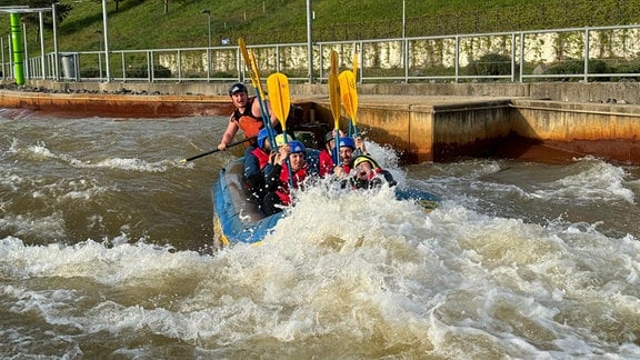 Ein Schlauchboot fährt in einem Wildwasserkanal durch Stromschnellen. Im Boot sitzen Menschen.