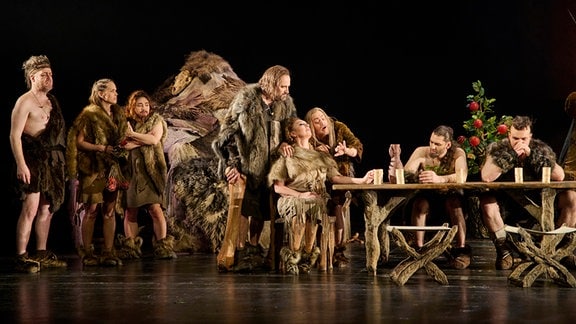 8 Menschen in Steinzeit-Kostümen sind auf der Bühne vor schwarzem Hintergrund. 