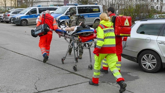 Medizinische Einsatzkräfte schieben einen Verletzten. Im Hintergrund Polizeiautos