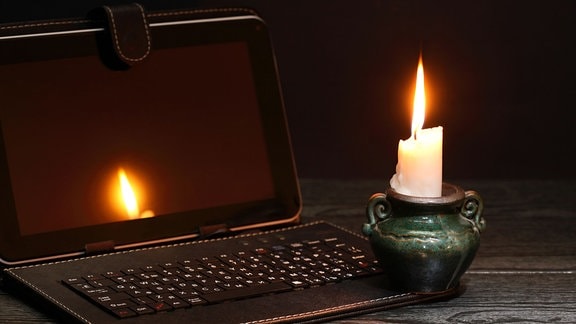 Eine brennende Kerze neben einem ausgeschaltetem Laptop in einem dunklen Raum.