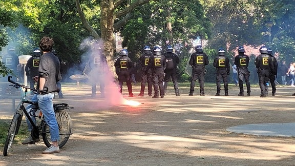 In einem Park stehen Polizisten in einer Reihe am weg. vor ihnen brennt ein roter Böller, der in ihre Richtung geschossen wurde. 