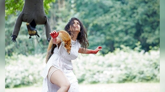 Eine Frau im Wald trägt ein weißes Kleid und einen ausgestopften Fuchs über der Schulter. Sie duckt sich angstvoll vor etwas nicht Sichtbarem über ihr.
