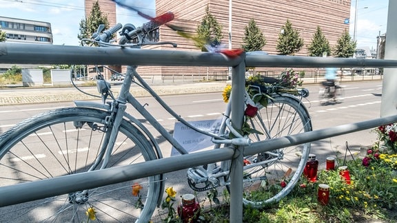 Ein komplett weiß lackiertes Fahrrad steht an einem Geländer neben einer dreispurigen Straße. Am Fahrrad sind Blumen angebracht. Daneben stehen Grabkerzen.