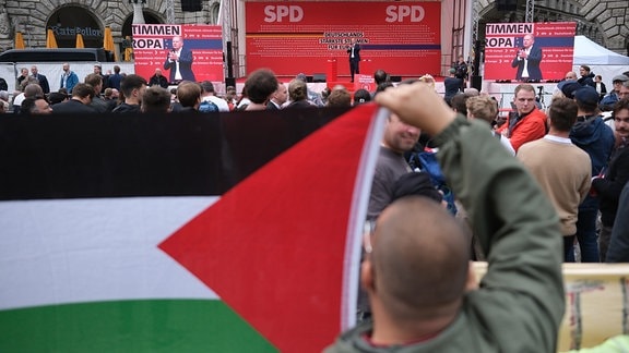 Olaf Scholz  spricht bei einem Wahlkampfauftritt, während Pro-Palästina-Aktivisten protestieren.