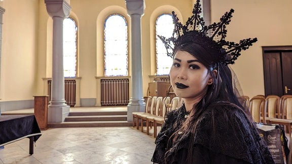 eine junge Frau mit langen schwarzen Kleid und schwarzer Kronenhaube sitzt in einer Trauerhalle