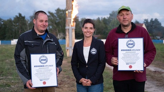 Robert Klauke (l) und Sebastian Müller, Organisatoren, stehen gemeinsam mit Eva Pastuszek, Rekord-Institut für Deutschland, vor einer 34 Meter hohen brennenden Fichte und halten eine Rekord-Urkunde.