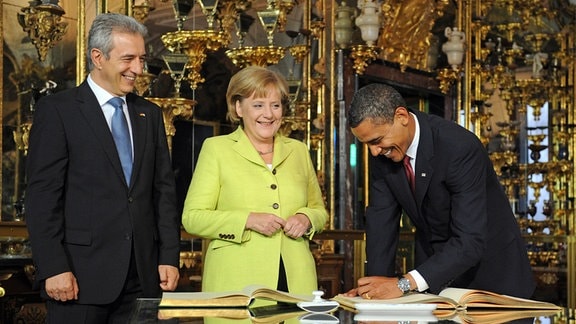 US-Präsident Barack Obama unterschreibt am 05.06.2009 die goldenen Bücher der Stadt Dresden und des Freistaates Sachsen im Preziosensaal des Dresdner Residenzschlosses. Links neben ihm stehen Bundeskanzlerin Angela Merkel (CDU) und der sächsische Ministerpräsident Stanislaw Tillich (CDU). 