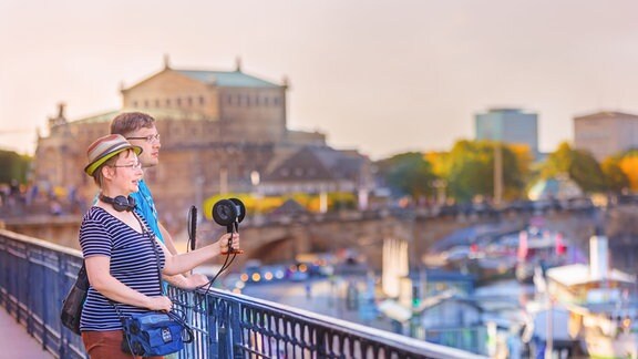 Eine Frau in einem gestreiften T-Shirt mit Hut auf dem Kopf und Mikrofon in der Hand steht neben einem Mann in einem blauen T-Shirt an einem Geländer. Sie blicken auf einen Fluss. Im Hintergrund sind Schiffe und die Silhouette einer Stadt zu sehen. 