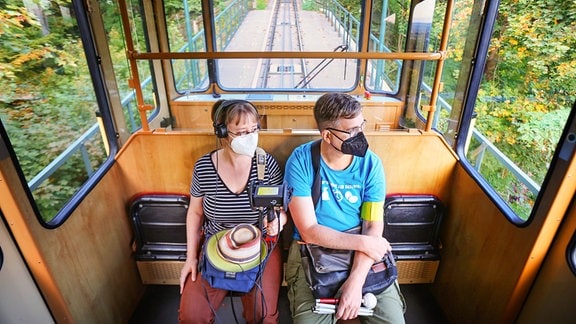 Eine Frau in einem gestreiften T-Shirt mit Kopfhörern um den Hals sitzt neben einem Mann in einem blauen T-Shirt mit Taststock in der Hand und gelber Armbinde. Sie fahren in einer Standseilbahn.