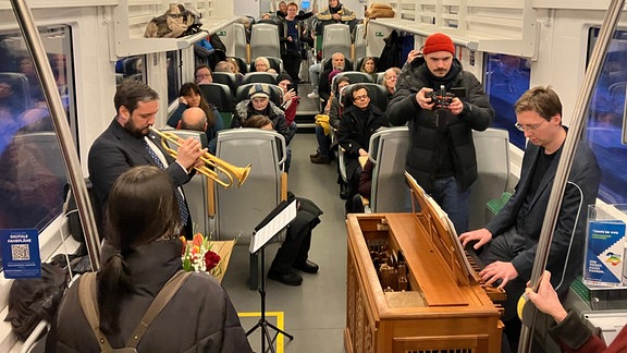 Vollbesetzter Zug, in dem ein Konzert mit einer Truhenorgel gespielt wird.