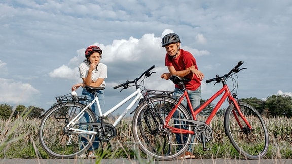 Tochter mit Vater, der sich auf ein Fahrrad stützt, während er auf einem landwirtschaftlichen Feld vor bewölktem Himmel steht.