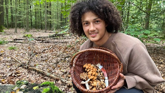 Ein junger Mann hält lächelnd einen Pilzkorb in den Händen