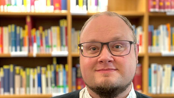 Friedemann Brause - ein junger Mann mit Brille steht vor einem Bücherregal und lächelt in die Kamera