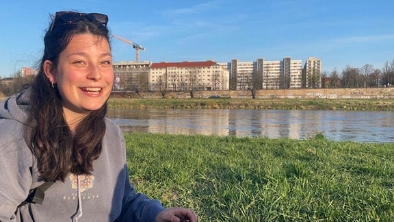 Eine junge Frau steht auf einer Wiese vor einem Fluss und lächelt in die Kamera