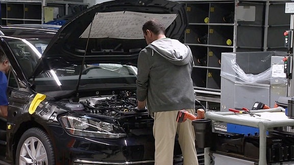 Ein Automonteur steht vor einem PKW mit geöffneter Motorhaube und schaut in den Motor.