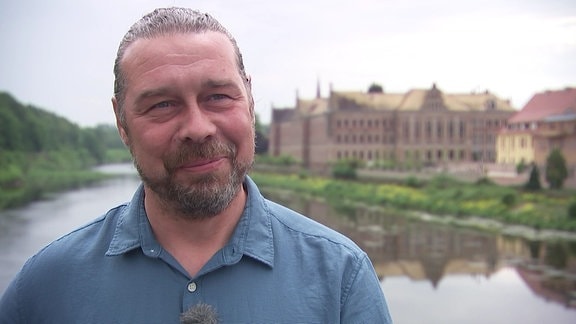 Ein Mann in einem blauen Hemd steh an einem Fluss, im Hintergrund ist ein großes Gebäude zu sehen.