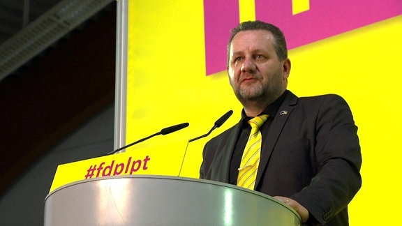 Mann mit dunklen Haaren und Vollbart in schwarzem Anzug mit gelbem Schlips vor gelb-rotem Hintergrund