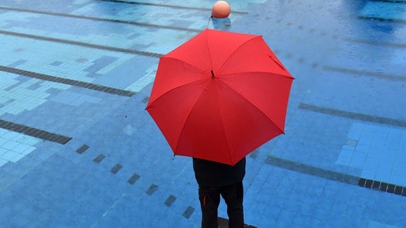 Ein Mitarbeiter eines Freibades steht bei Regenwetter mit einem Schirm am Sprungbecken.