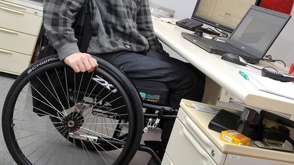 Ein Mann sitzt in einem Rollstuhl an seinem Arbeitsplatz am Schreibtisch. 