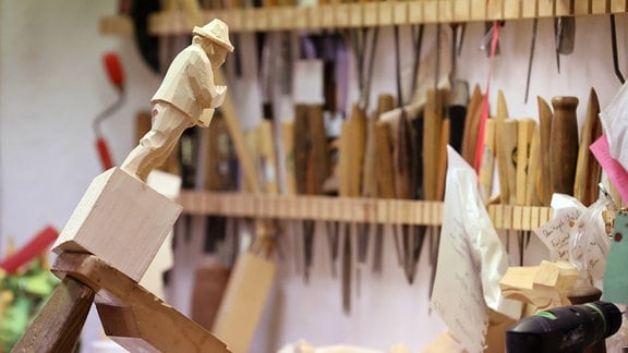 In einer Schauwerkstatt ist eine Figur eingespannt und wartet darauf, fertig geschnitzt zu werden, im Hintergrund sieht man die unterschiedlichen Werkzeuge.