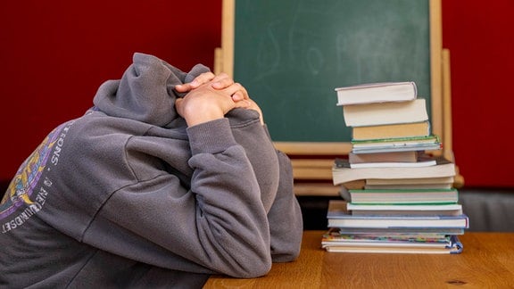 Eine Schülerin sitzt vor einem Stapel Büchern und hat trotzig die Kapuze ihres Pullovers über den Kopf gezogen. In Nordrhein-Westfalen haben 2022 deutlich mehr Jugendliche die Schule ohne Hauptschulabschluss verlassen.