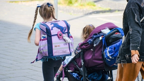 Kleines Mädchen geht mit ihrem Ranzen auf dem Rücken gemeinsam mit  einer Erwachsen Person die einen Kinderwagen mit einem Kleinkind schiebt einen Fußweg entlang.