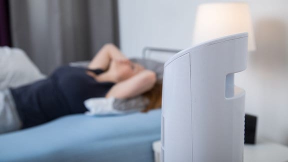 Eine junge Frau liegt im Bett, davor steht ein Ventilator