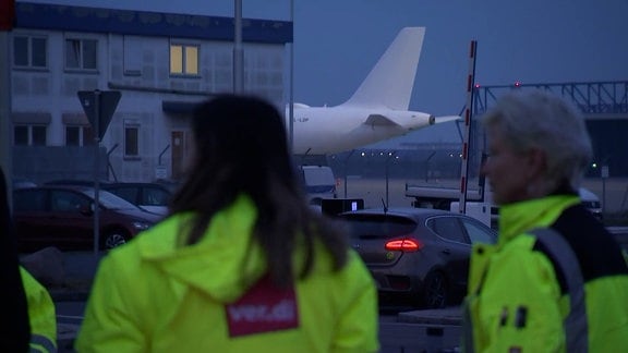 Zwei Frauen in Verdi-Westen gekleidet im Vordergrund. Im Hintergrund ein Flugzeug. 
