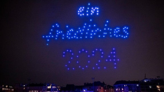 Leuchtende Drohnen fliegen bei einer Drohnenshow über einem Stadion und formen dabei am Himmel den Schriftzug "ein friedliches 2024". 
