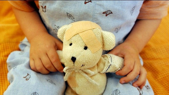 Ein Kind hält einen Teddy mit Pflaster auf dem Kopf und verbundenem Arm