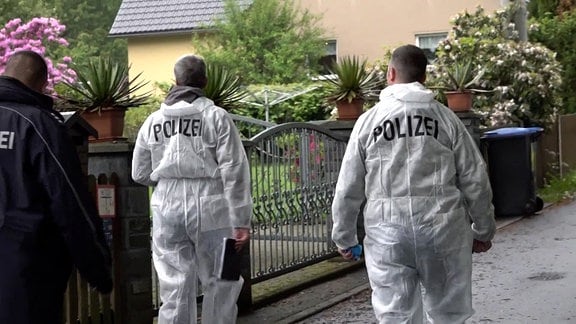 Zwei Personen von der Spurensicherung in weißen Schutzanzügen und ein Polizist vor einem Haus mit blühendem Vorgarten