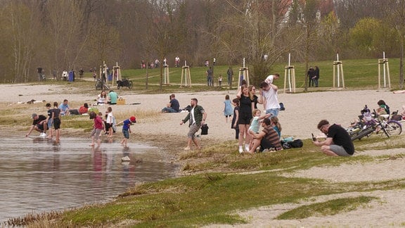 Am Sandstrand des Cospudener Sees sitzen Menschen, einige stehen mit den Füßen im Wasser. 