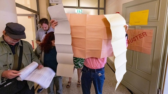 Wähler mit drei ausgebreiteten Stimmzetteln vor einem Wahlbüro