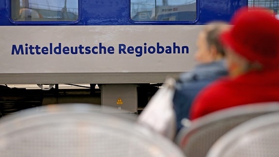 Zwei Reisende sitzen vor einem Zug mit der Aufschrift "Mitteldeutsche Regiobahn".