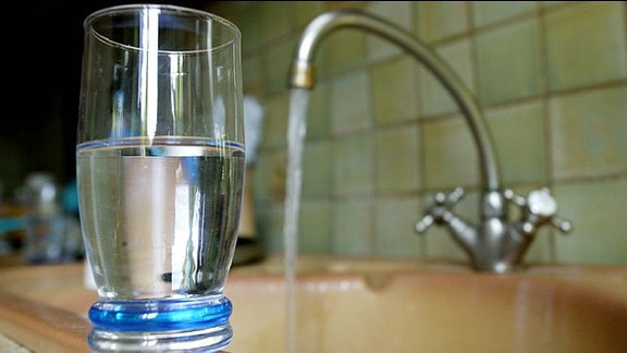 Ein Glas mit Wasser und im Hintergrund ein Wasserhahn.