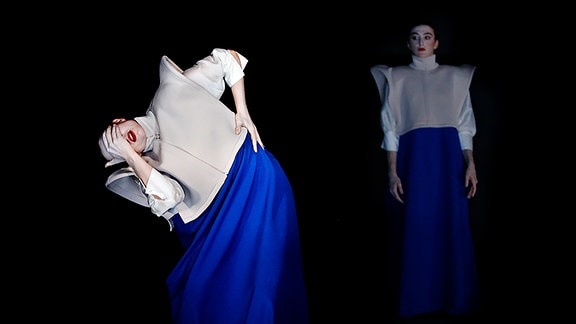 Szene auf einer Theaterbühne auf der sich zwei weiß geschminkte Schauspielerinnen in hellen ausladenden Oberteilen und blauen weiten Röcken befinden.