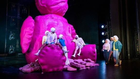 Auf einer Theaterbühgne ist ein großes rosafarbenes Tier zu sehen, Menschen sitzen auf seinen Füßen. 