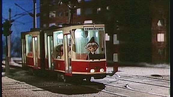 Die Sandmännchenpuppe sitzt bei Abendstimmung im Führerstand einer roten Tatra-Straßenbahn